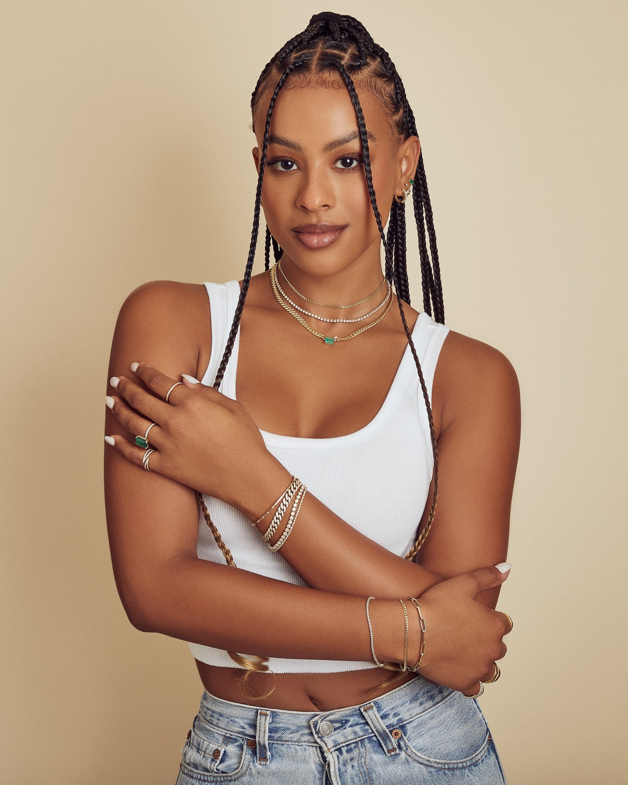 Woman with braided hair wearing a white tank top and jeans against a beige background with lots of gold and emerald jewelry