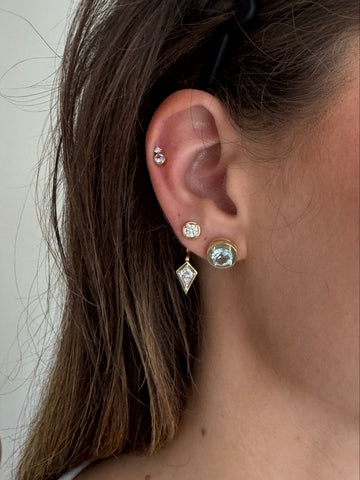 A close-up of a person's ear wearing multiple stud earrings, including a gold-toned round stud with aquamarine, a small gemstone stud with pink sapphire and diamond, a kite-shaped  double stud, with brown hair framing the ear.